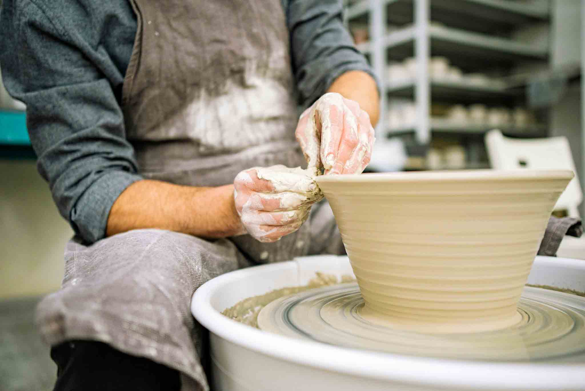 Potter shaping a clay pot on a spinning pottery wheel with hands covered in wet clay.